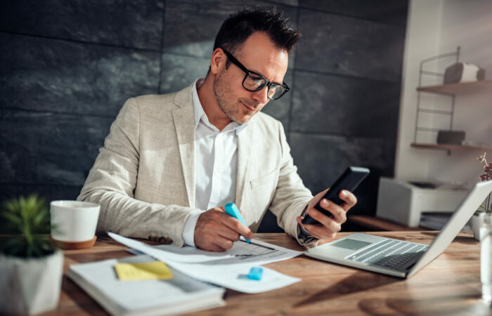 Profissional usando marcador em documentos, sentado à mesa de escritório com notebook e segurando celular para importar XML