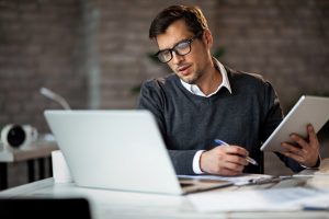 Homem segurando tablet com uma mão, caneta com a outra e olhando para a tela de um notebook, que simbolizando codigo cest