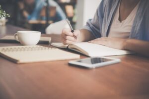 Sobre uma mesa estão uma xícara, um caderno aberto, um celular e um livro de anotações no qual uma mulher está anotando rendimentos tributáveis