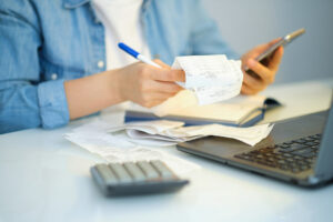 Pessoa segurando celular com uma mão e notas fiscais com caneta na outra mão, a pessoa está sentada à mesa de escritório. Sobre a mesa estão um notebook, um caderno aberto, mais notas fiscais e uma calculadora. Simbolizando o cálculo de impostos sobre vendas