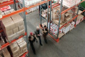 Três trabalhadores andando dentro de um depósito com grandes prateleiras e inúmeras caixas e embalagens, simbolizando classificação fiscal de mercadorias