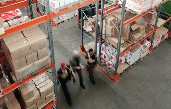 Três trabalhadores andando dentro de um depósito com grandes prateleiras e inúmeras caixas e embalagens, simbolizando classificação fiscal de mercadorias