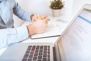 Simbolizando Sped Fiscal: Homem fazendo anotações em caderno ou agenda aberta, que está sobre uma mesa de escritório. Sobre a mesma mesa também está um vasinho de plantas, um celular e um notebook