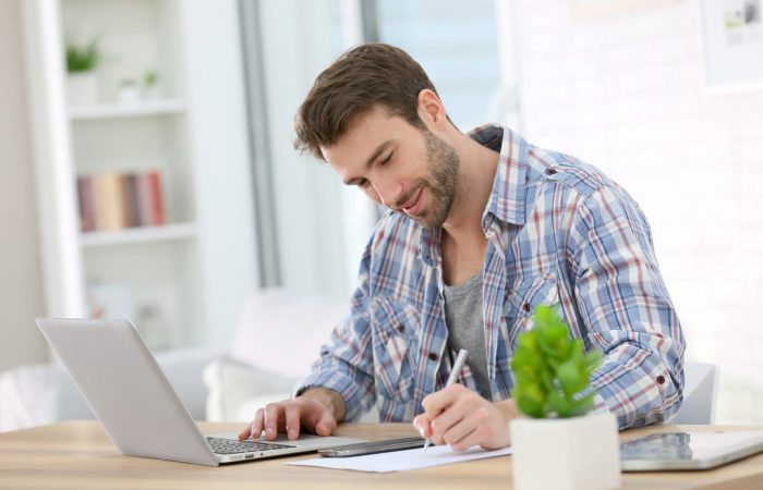 Simbolizando como emitir nota fiscal avulsa: Homem com expressão feliz usando notebook e escrevendo em folha de sulfite com a outra mão