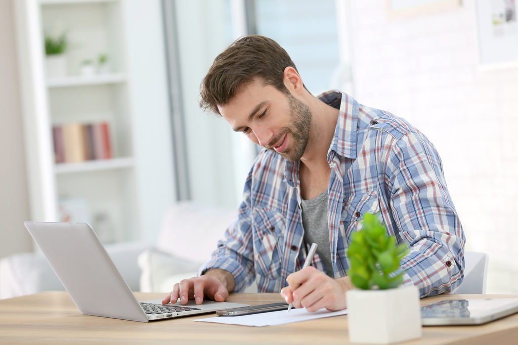 Simbolizando como emitir nota fiscal avulsa: Homem com expressão feliz usando notebook e escrevendo em folha de sulfite com a outra mão