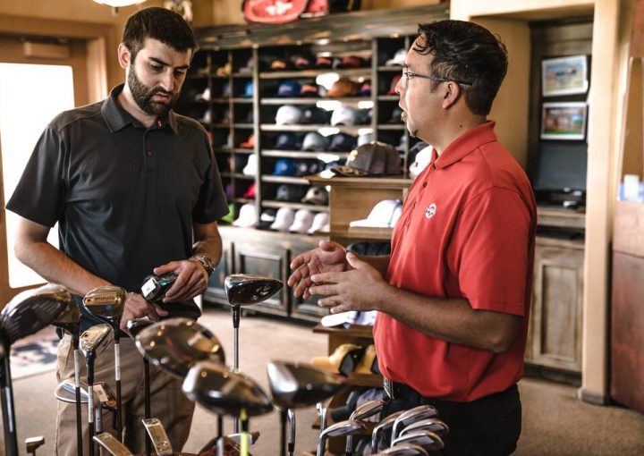 Atendente especialista mostrando tacos de golf para cliente e fazendo explicação dentro de loja de bonés e esportes