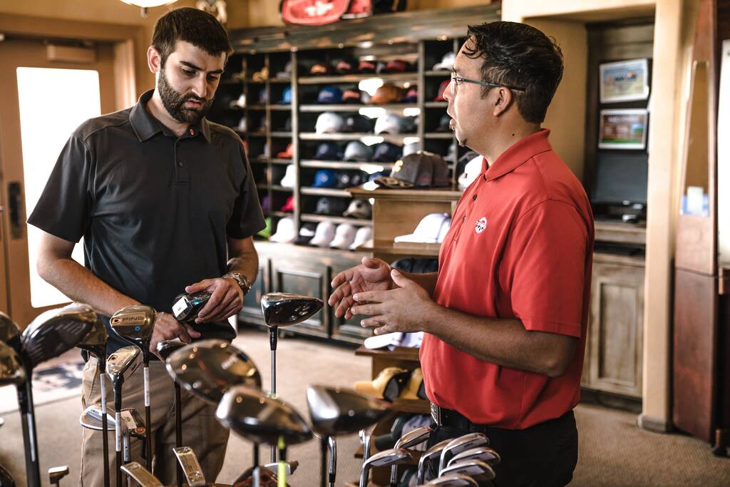 Atendente especialista mostrando tacos de golf para cliente e fazendo explicação dentro de loja de bonés e esportes