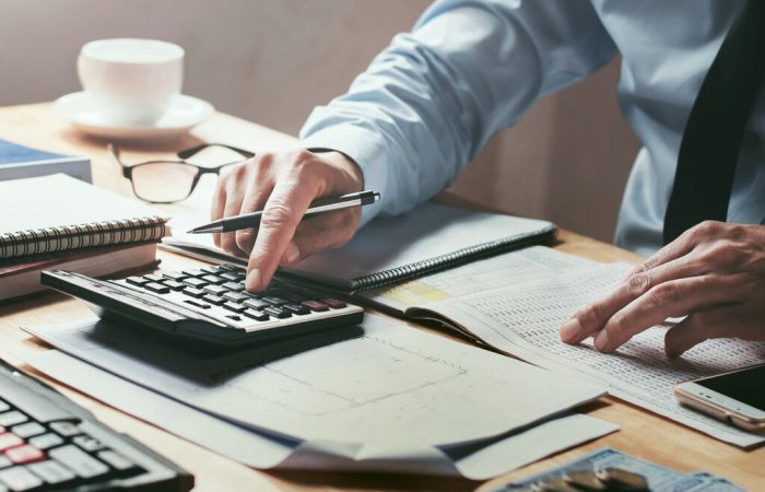 Simbolizando livro caixa, pessoa segurando caneta e usando calculadora, que está sobre a mesa de um escritório que contém também: documentos, uma xícara e óculos de grau