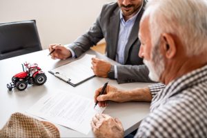 Idoso assinando documento ao lado de homem vestido com terno cinza. Na mesa, um trator em miniatura vermelho.