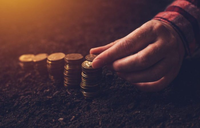 Detalhe de mão posicionando diversas moedas em pilhas de tamanhos diferentes, do menor para o maior, em formato de gráfico de crescimento, sob terra de plantio.