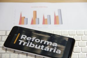 Celular com imagem da reforma tributária na tela, em cima de teclado e gráficos de crescimento econômico.