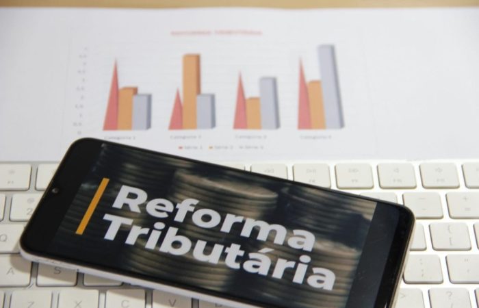Celular com imagem da reforma tributária na tela, em cima de teclado e gráficos de crescimento econômico.
